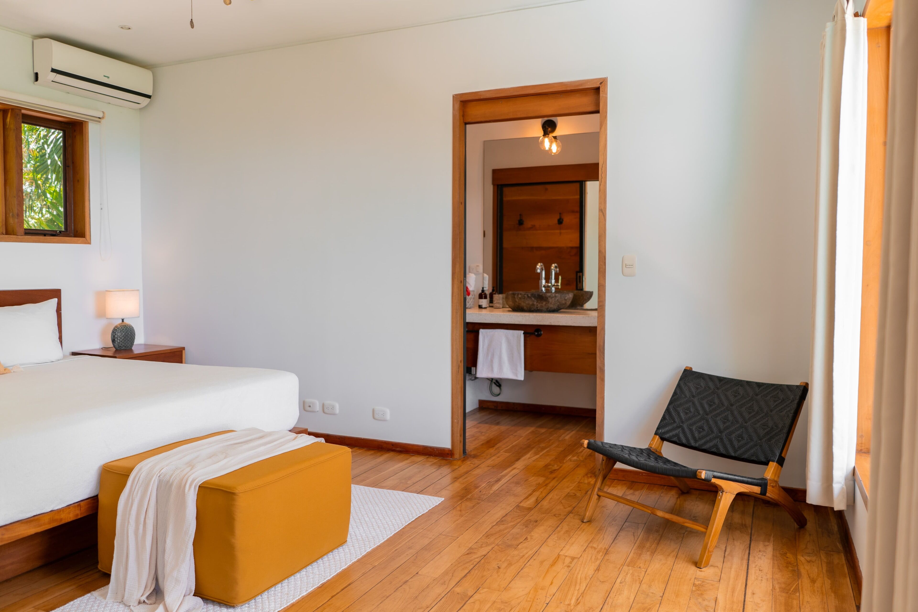 A bedroom with a bed, a wooden bench, a chair, a nightstand, and a window with blinds.