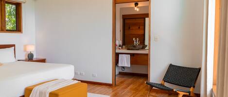 A bedroom with a bed, a wooden bench, a chair, a nightstand, and a window with blinds.
