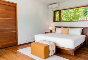 A bedroom with a wooden bed frame, white bedding, a nightstand with a lamp, and a window with a view of trees.