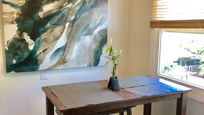 Ein Zimmer mit einem Holztisch, zwei Hocker, einem großen abstrakten Gemälde an der Wand und einem Fenster mit Jalousien.