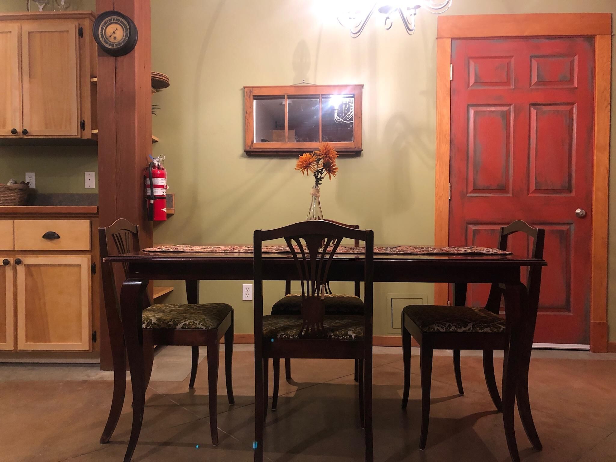 Un coin repas avec une table en bois, quatre chaises, un vase de fleurs, une horloge et une porte rouge.