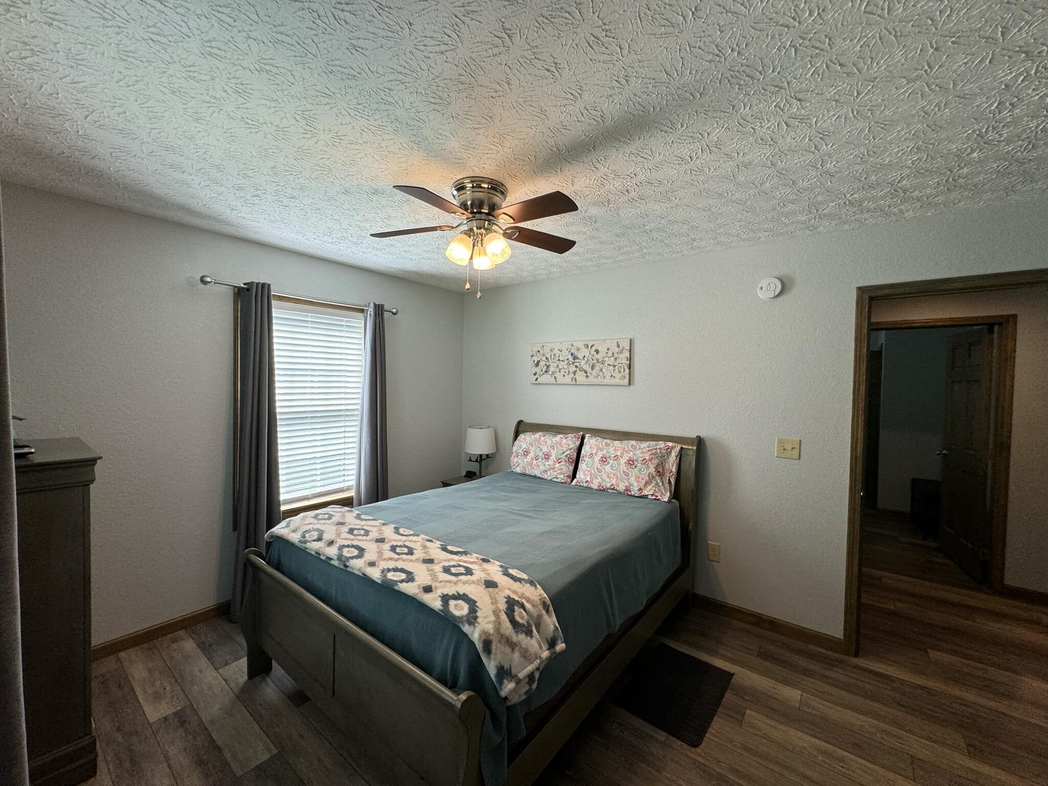 Un dormitorio con cama, ventilador de techo, ventana con cortinas, mesita de noche con lámpara y puerta.