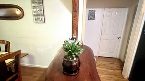 Uma sala de jantar com uma mesa de madeira, cadeiras e uma planta em vaso. Há dois avisos na parede, um com uma citação e outro com uma mensagem.