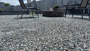 Área externa com assentos, duas cadeiras de metal e um pequeno braseiro sobre piso de brita, cercada por um muro de madeira.