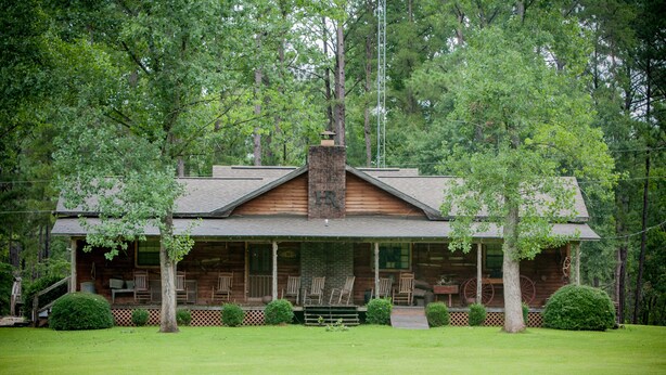 A log cabin with a porch, surrounded by trees and a well-maintained lawn.