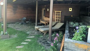 Overdekt terras met een houten bank, een tafel en een stoel. Er is een tuin met bloemen en een houten pergola.