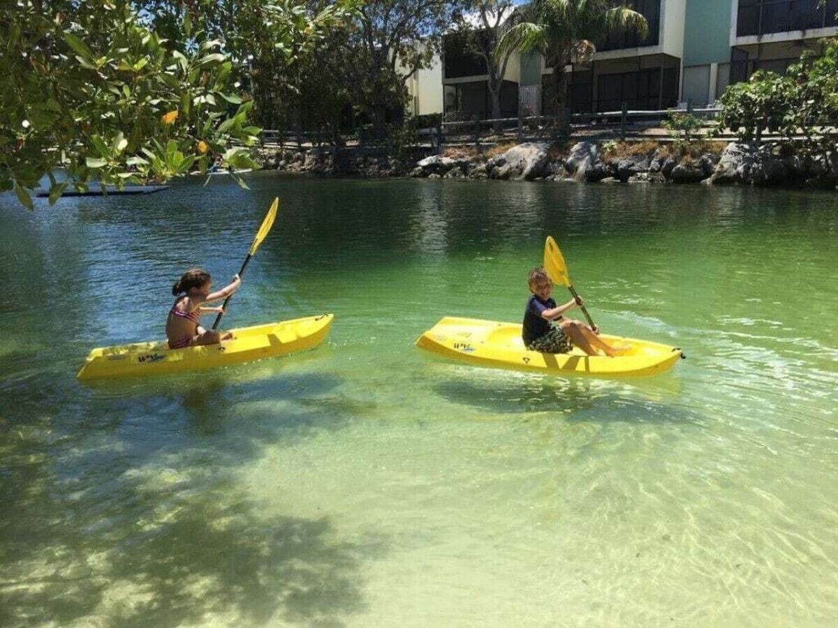 Two people kayaking on a calm body of water surrounded by greenery and buildings.