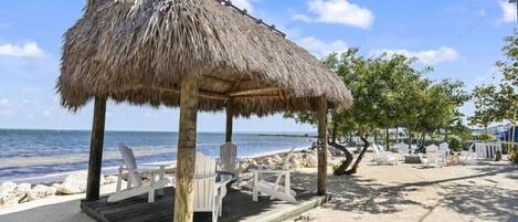 A beachside gazebo with a thatched roof, white chairs, and a wooden deck.