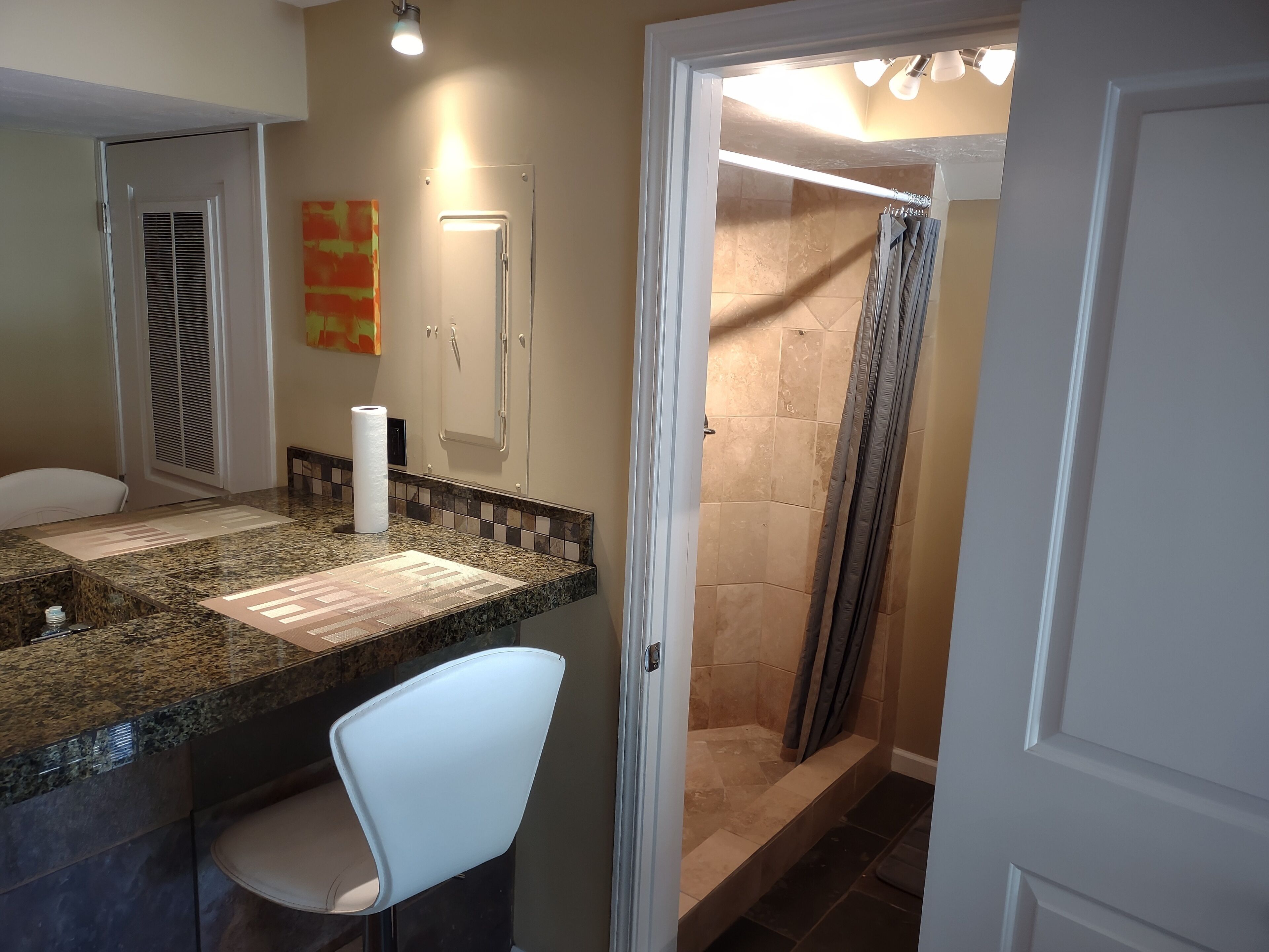 A bathroom with a granite countertop, a white chair, a shower with a glass door, and a towel dispenser.