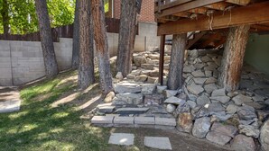 A stone pathway leading to a wooden deck under a rustic structure, surrounded by trees and a brick wall.