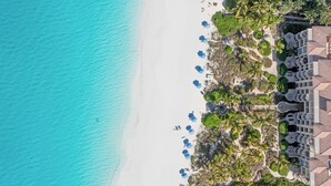 Vue aérienne d’un complexe hôtelier en bord de mer, avec une plage de sable blanc, une eau turquoise et limpide, et une rangée de parasols bleus.