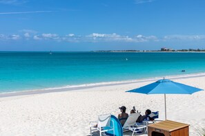 A beach with white sand, a clear blue sea, and a few people relaxing under a blue umbrella.