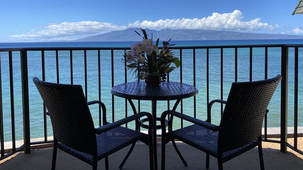Un balcón con dos sillas de mimbre y una pequeña mesa redonda con un arreglo floral, con vista a un mar azul y una montaña distante.