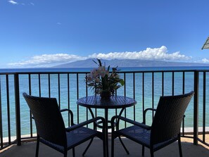 A balcony with two wicker chairs and a small round table with a flower arrangement, overlooking a clear blue sea and a distant mountain.