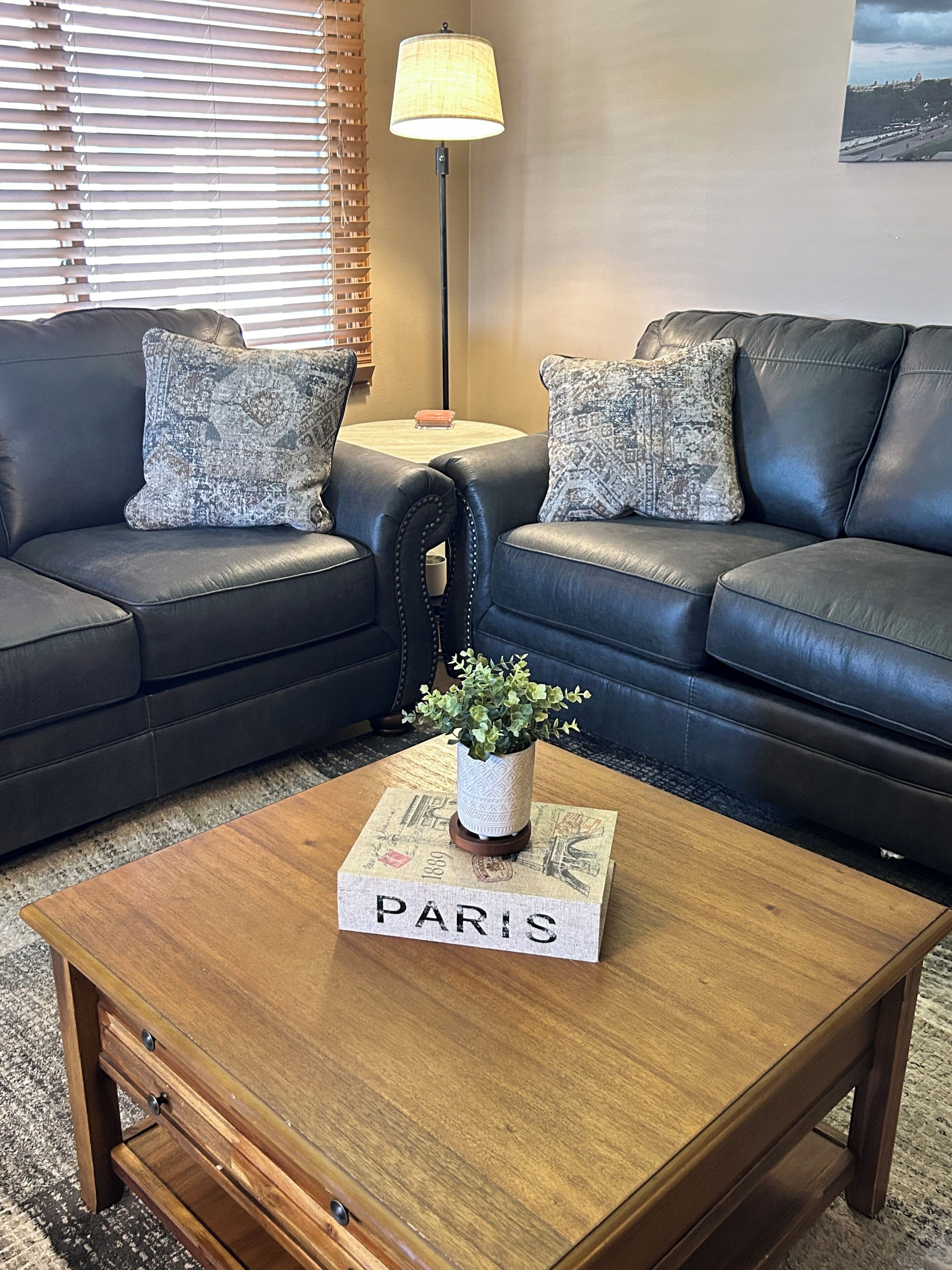 A living room with two black leather sofas, a wooden coffee table with a 'PARIS' placard, a small potted plant, and a lamp on a side table.