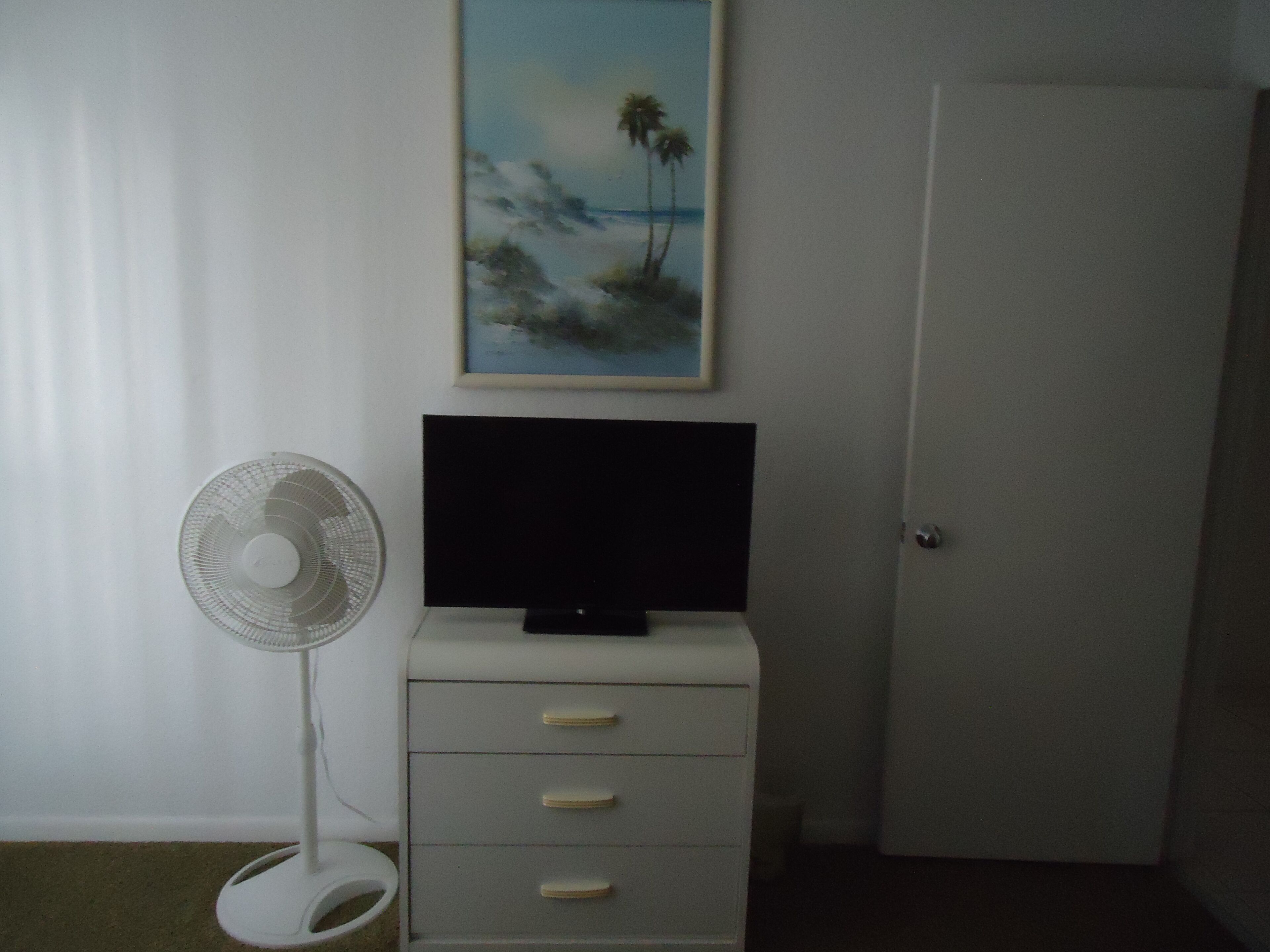 Una habitación con un aparador blanco, un televisor de pantalla plana encima, un ventilador de pie y una puerta.