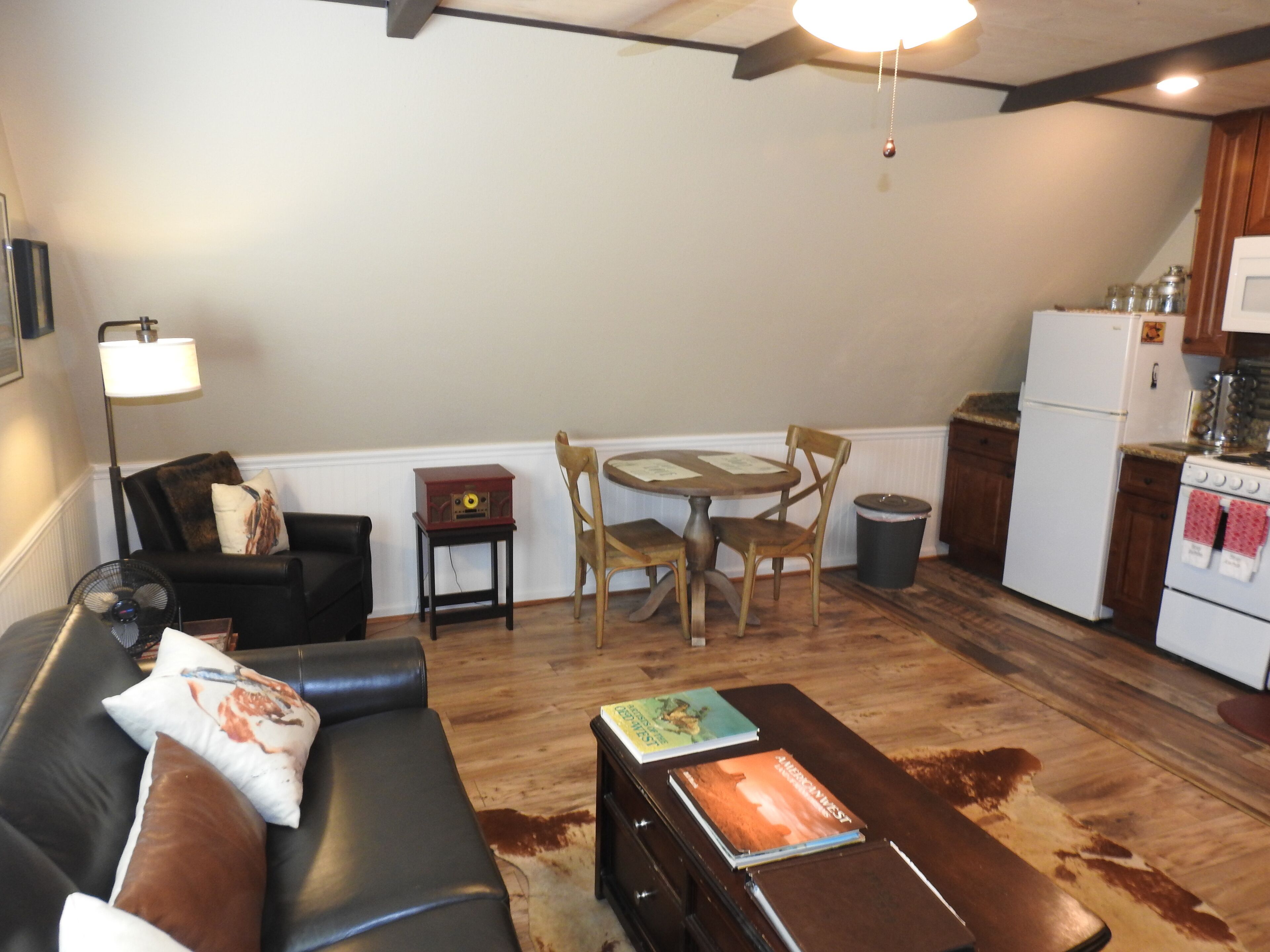 Una sala de estar con un sofá, una mesa de centro con libros, una mesa de comedor con sillas, una radio pequeña y una zona de cocina con refrigerador y estufa.