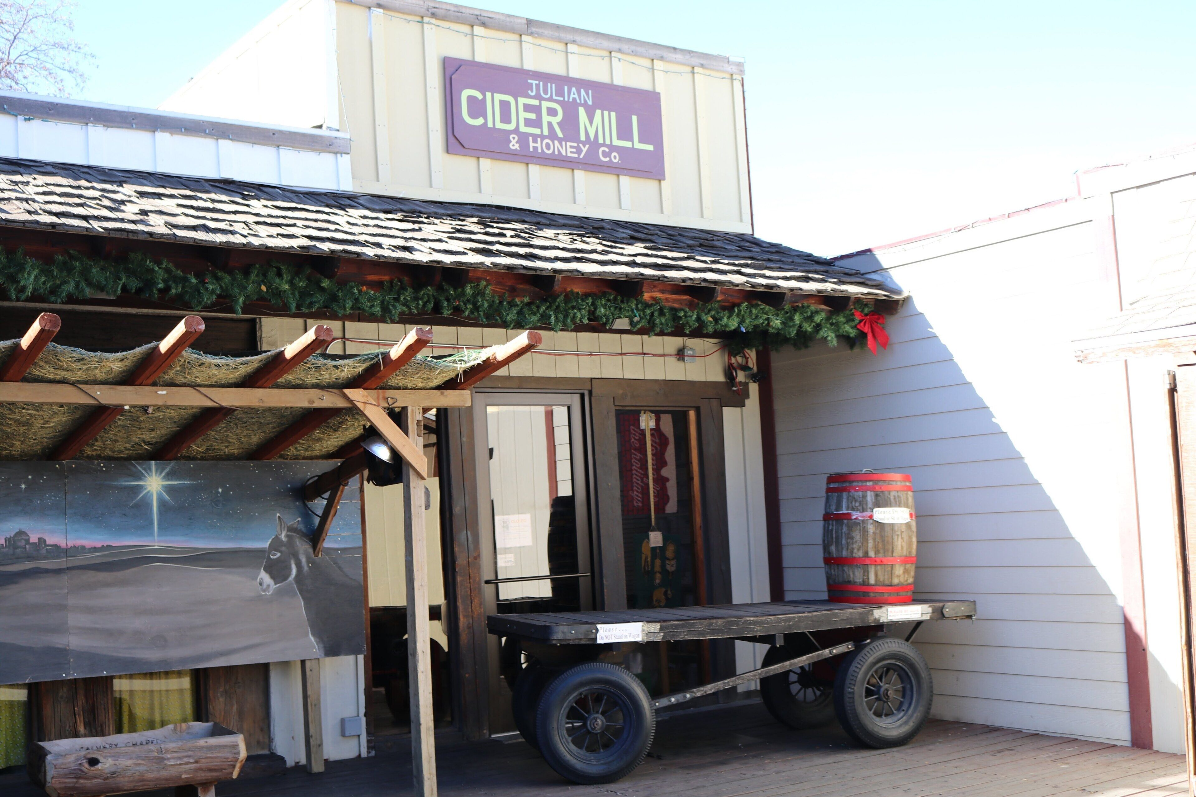 A building with a sign reading "Julian Cider Mill & Honey Co." and a wooden cart with a barrel next to it.