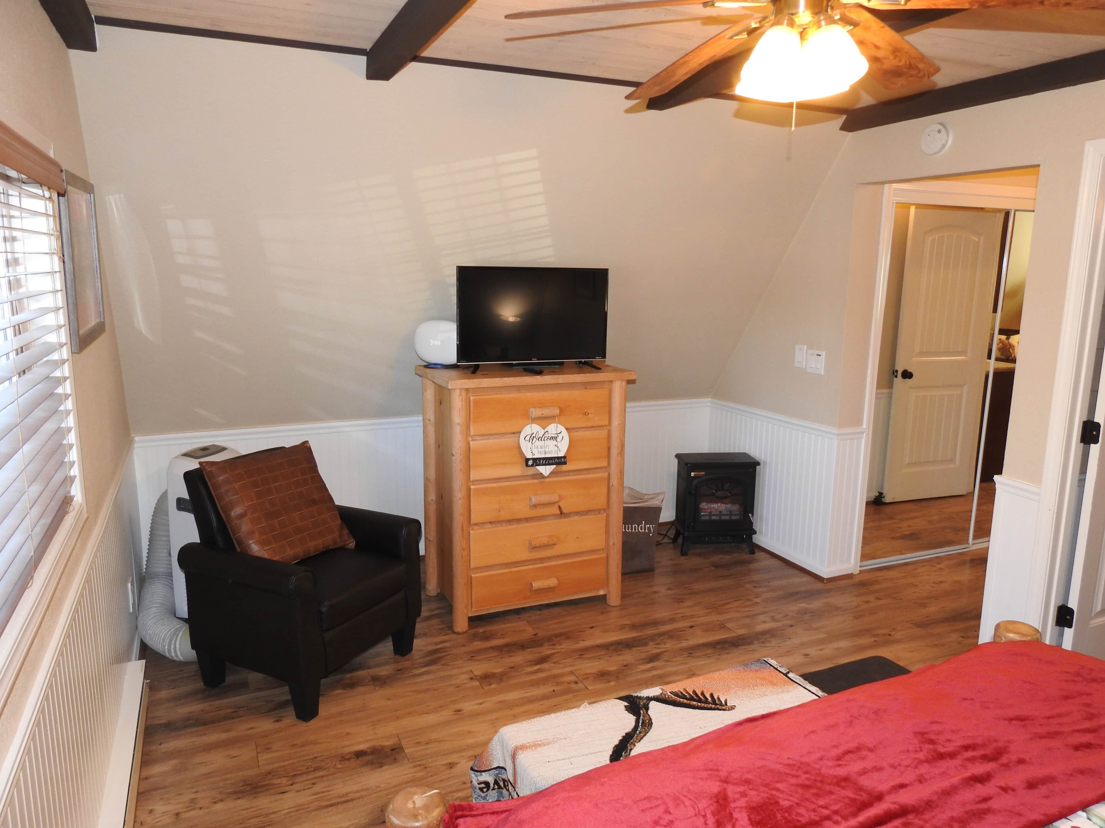 A bedroom with a wooden dresser, a television on top, a black chair, a red blanket, and a wooden ceiling fan.