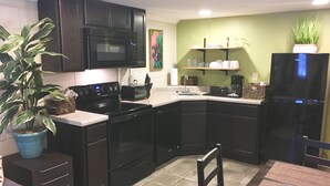 Eine kleine Küche mit dunklen Schränken, einer Mikrowelle, einem Herd und einem Kühlschrank. Es gibt ein Waschbecken, einen Esstisch mit Stühlen und eine Topfpflanze.