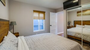 Een slaapkamer met een bed, een nachtkastje met een lamp, een raam met jaloezieën, en een deur die naar een andere kamer leidt.