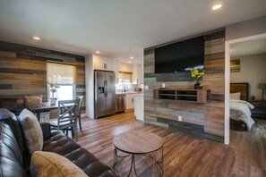 A modern living room with a leather sofa, a round coffee table, and a dining area with a wooden table and chairs. A kitchen with a refrigerator and a flat-screen TV is visible in the background. - Modern Renovated Bayside condo-808 (San Diego)