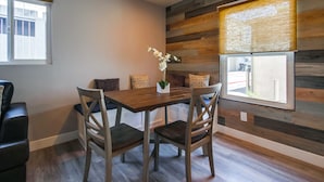 Een eethoek met een houten tafel en stoelen, een vaas met bloemen en uitzicht door een raam met jaloezieën.