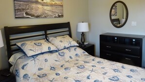 A bedroom with a bed, a nightstand, a lamp, a dresser, a mirror, and a painting of a beach scene.