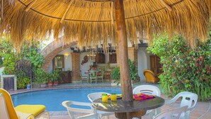 An outdoor pool area with a thatched umbrella, white plastic chairs, and a dining table with chairs.