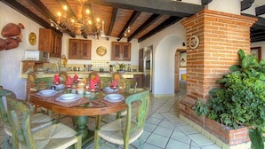 A dining area with a wooden table set for four, surrounded by green chairs. A brick fireplace is visible to the right, and a kitchen area with wooden cabinets and a hanging light fixture is in the background.