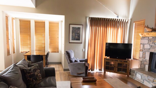A living room with a sofa, a television, a stone fireplace, and wooden blinds.
