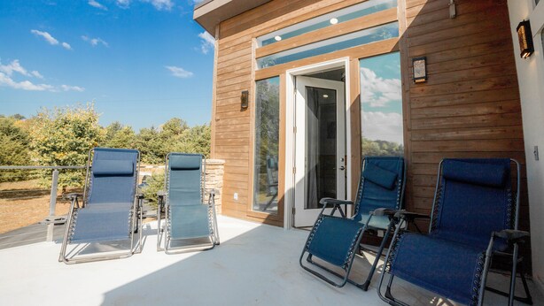 A wooden building with a glass door and windows, a covered patio with four blue lounge chairs, and a clear blue sky. - Fox Den at Fox Hollow (Aldrich)