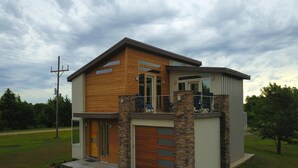 A modern two-story house with a wooden facade, a balcony with outdoor seating, and a garage. - Fox Den at Fox Hollow (Aldrich)