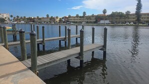 A waterfront dock with a wooden walkway leading into calm water, surrounded by buildings and palm trees.