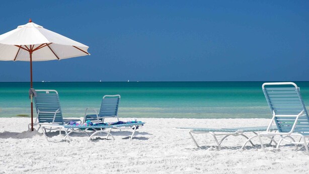A beach with white sand, clear turquoise water, and a white umbrella providing shade. There are several empty lounge chairs and a small table with a towel.