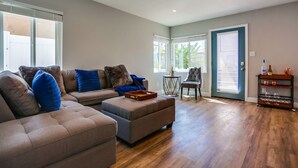 A spacious living room with a large sectional sofa, a small ottoman, and a wooden coffee table. There is a dining area with a chair and a small table. The room has hardwood flooring and large windows with blinds. - Renovated Bay Side Condo - 812 (San Diego)