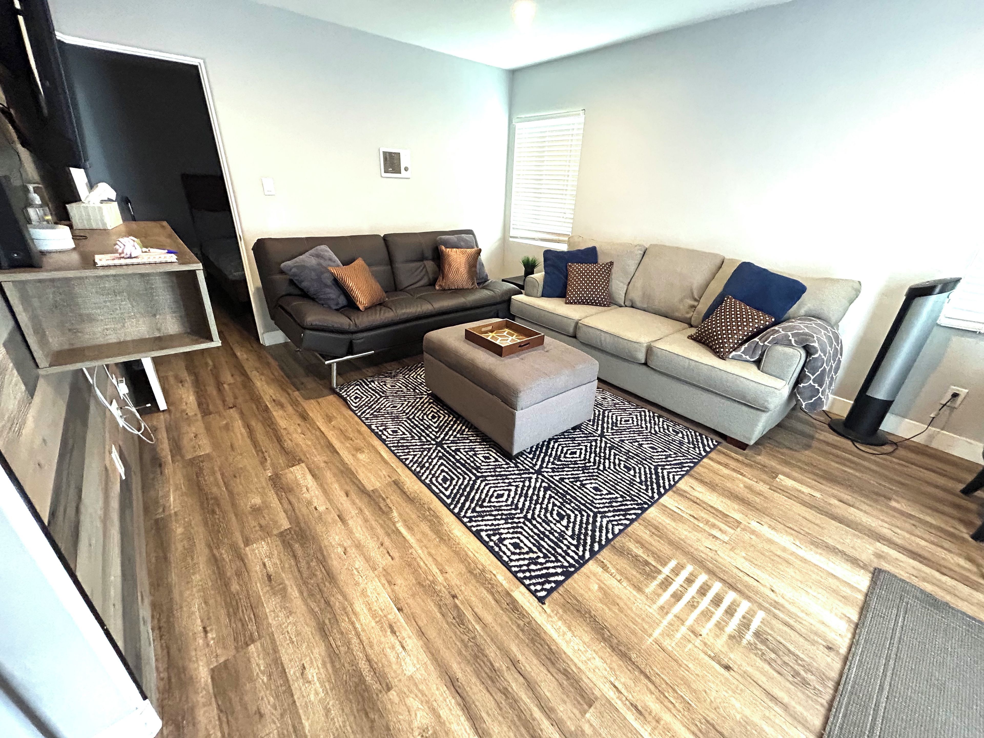 Un salon moderne avec un canapé, un pouf et une table basse. On y trouve également un téléviseur et une fenêtre avec des stores.