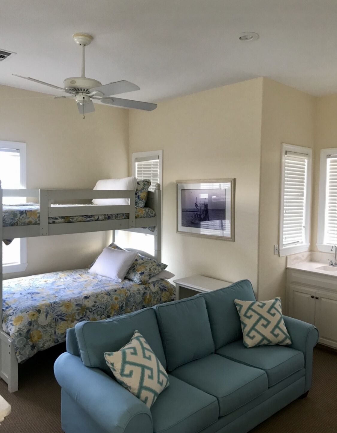 A room with a bunk bed, a sofa, a ceiling fan, and a framed picture on the wall.