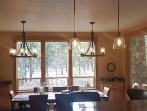 A dining area with a wooden table and chairs, pendant lights, and a view of trees through large windows.