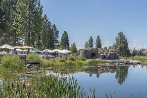 A lakeside area with outdoor seating under umbrellas, a rocky waterfall structure, and surrounding trees.