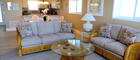 A living room with two sofas, a glass coffee table, wicker chairs, and a kitchen in the background.