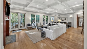 Un salon spacieux avec un canapé blanc, une table basse et un lustre. De grandes fenêtres et une porte donnent accès à un espace extérieur.