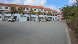 A multi-story building with a red-tiled roof, white balconies, and a paved parking area.