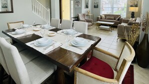 A dining area with a wooden table set for four, surrounded by upholstered chairs. There is a staircase leading to another level, a sofa set, and a large window with blinds. - Florida Winter Escape 🌴 6BR Private Pool Snowbird + Family Visits (Kissimmee)