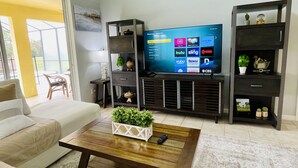 A living room with a large flat-screen TV, a sofa, a coffee table with a plant, and a bookshelf. - Florida Winter Escape 🌴 6BR Private Pool Snowbird + Family Visits (Kissimmee)