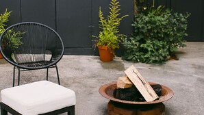 Un patio con dos sillas de metal negro y dos bancos blancos acolchados, un pequeño hogar con leña y plantas en macetas.