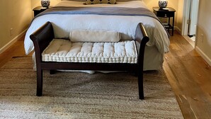 Un dormitorio con una cama grande, dos mesitas de noche con lámparas, una lámpara colgante y un banco frente a la cama.