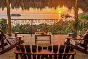 Une terrasse offrant une vue sur le coucher de soleil sur la mer, des meubles en bois et une table avec une bouteille et des verres.