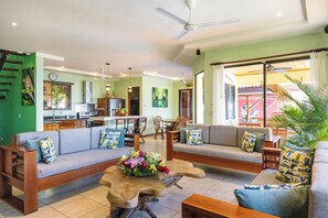 A spacious living area with a sofa set, a wooden coffee table with flowers, and a dining area in the background.