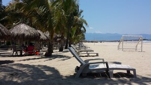 Una playa con sombrillas de paja, sillas de descanso y una portería de fútbol.